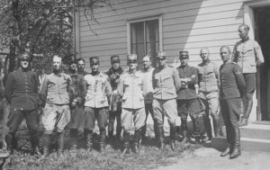 Motiv: Grupporträtt av personal vid Flygkompaniets flygskjutskola framför kavaljersflygeln på flygfältet vid Stora Lund, Hästholmen, 1923. Vid besök av chefen för Fälttelegrafkåren, Conrad Ericsson. Tillhör Gamla Malmen-samlingen. GM 1507. Ur Flygvapenmuseums bildarkiv.