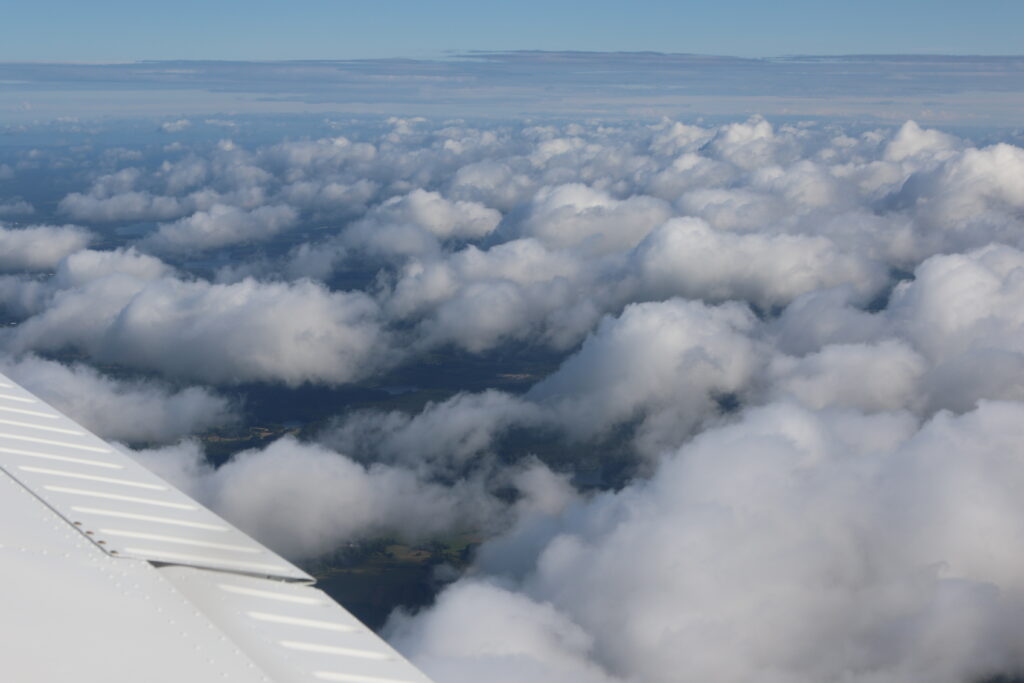 Flygfoto taget strax ovanför topparna av ett lager av låga cumulusmoln. Täckningsgraden är c:a 50%, så marken syns mellan molnen nära flygplanet.
