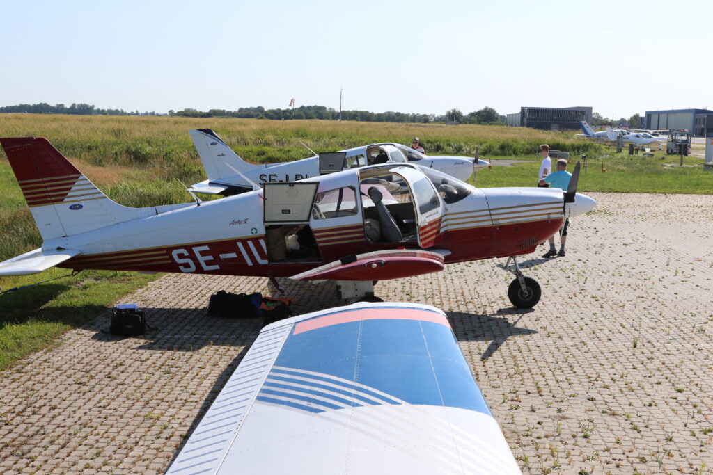 Två små enmotoriga propellerflygplan parkerade bredvid varandra i utkanten av en stenlagd platta med stjärtarna ut över gräset. Fotot är taget från taket av ett av planen mot de andra två. Dörrar och lastluckor är öppna, bagage ligger på marken och besättningarna håller på att göra planen redo för start.