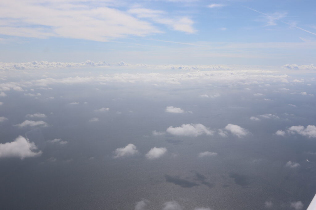 Flygbild tagen över vatten. Låga moln är vitt spridda nära flygplanet, men ser ut att vara tätare längre fram.