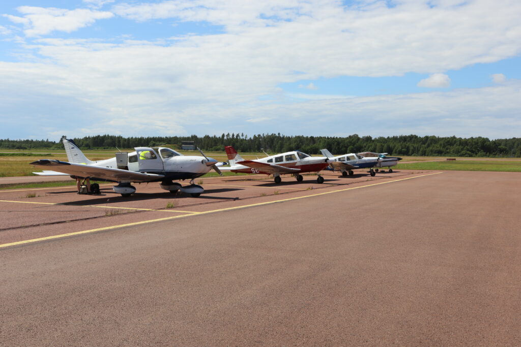 Tre PA-28:or parkerade i parkeringsrutor vid kanten av en asfaltsplatta under en himmel med tunna sommarmoln.